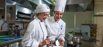 En una cocina profesional, dos chefs se encuentran trabajando juntas. Ambas llevan chaquetas de chef y gorros blancos. Una de ellas, con una sonrisa, utiliza un mortero para preparar ingredientes, mientras que la otra observa atentamente, proporcionando orientación.