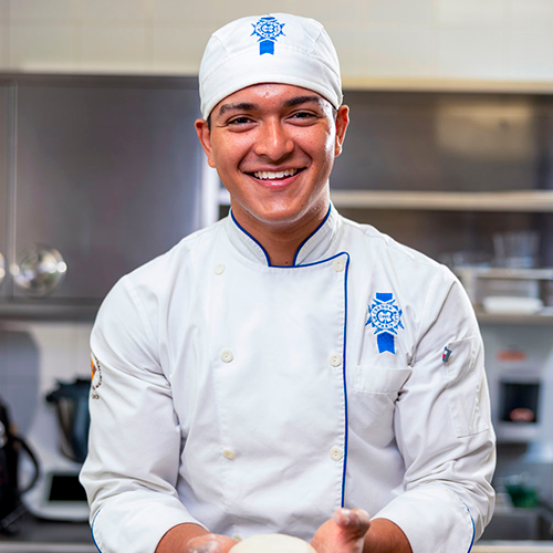 Joven chef sonriendo con uniforme blanco y gorro del instituto culinario Le Cordon Bleu, en una cocina profesional con utensilios de fondo, listo para comenzar una preparación.