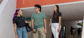 Tres estudiantes universitarios sonrientes conversan mientras caminan por el campus, reflejando un ambiente dinámico y amigable.