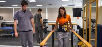 Un estudiante de Terapia Física con uniforme clínico asiste a una paciente que realiza ejercicios de marcha con el apoyo de barras paralelas en un gimnasio o clínica de rehabilitación. Otros estudiantes y equipos de terapia se ven en el fondo. La escena representa la práctica y el entrenamiento en Terapia Física.