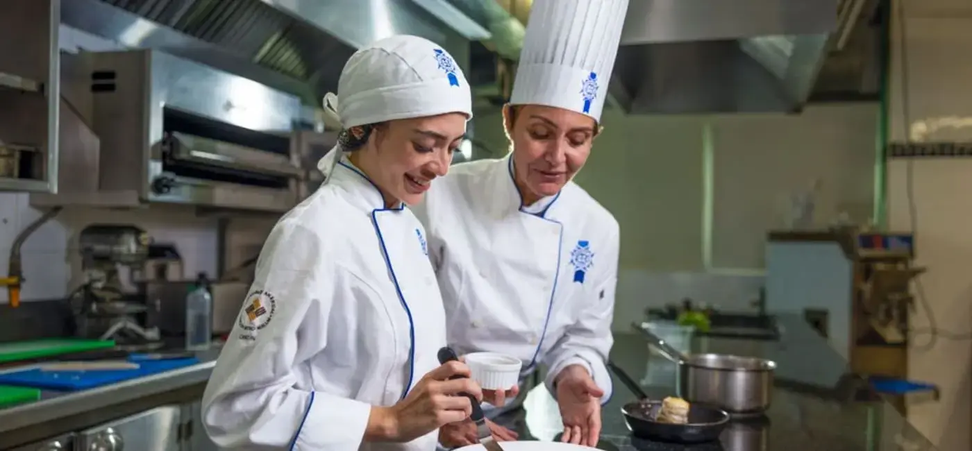 Una estudiante y una chef instructora con uniformes de Le Cordon Bleu colaboran en el emplatado de un platillo dentro de una cocina industrial profesional.