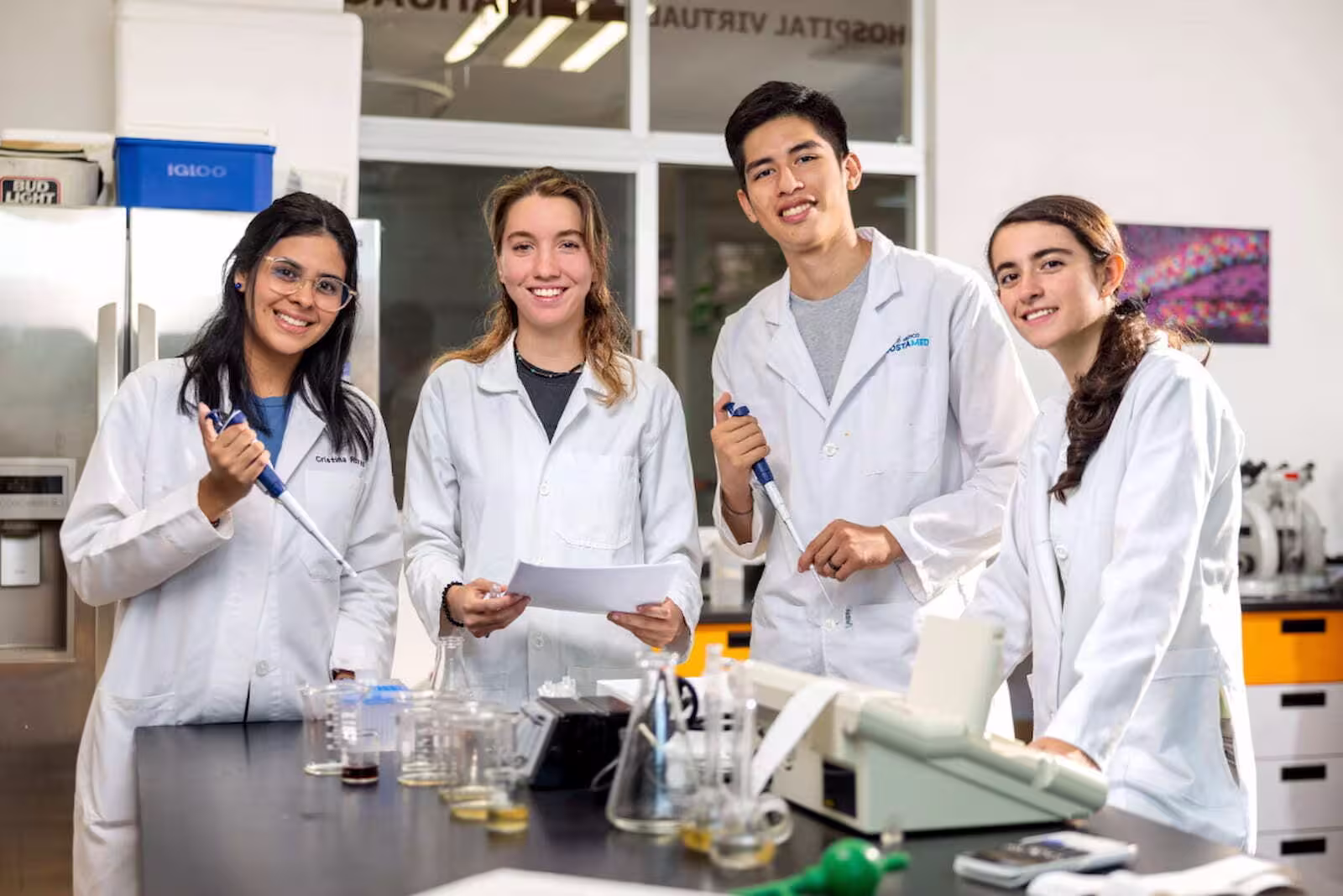Grupo de cuatro estudiantes de medicina y ciencias de la salud con batas blancas trabajando con pipetas y equipo de laboratorio en una mesa de prácticas.