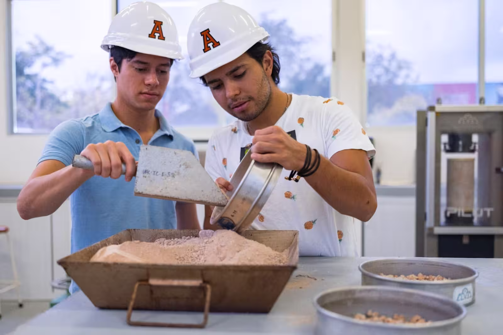 Dos estudiantes de ingeniería con cascos blancos de seguridad realizando una práctica de tamizado de materiales y suelos en un laboratorio universitario.