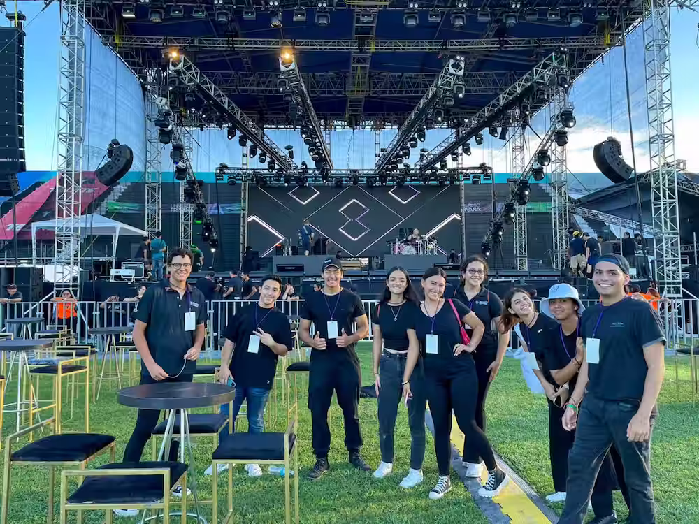 Grupo de estudiantes universitarios con gafetes de organización posando sonrientes frente a un gran escenario de conciertos al aire libre.