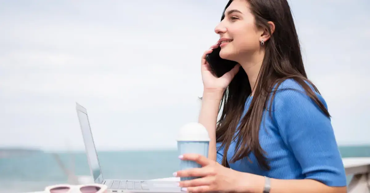 Una estudiante sonriente sentada al aire libre frente al mar, hablando por teléfono móvil mientras tiene una computadora portátil abierta y sostiene un café.