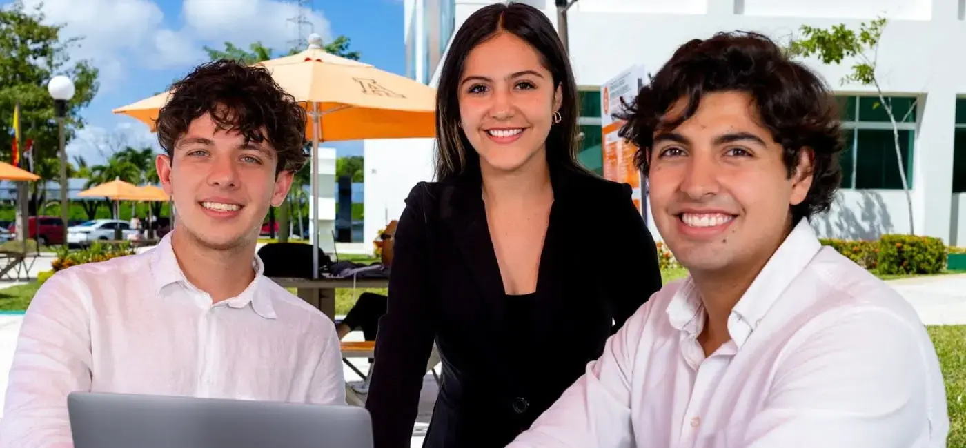 Tres estudiantes universitarios con vestimenta casual de negocios sonríen a la cámara mientras comparten una laptop al aire libre en el campus, destacando una zona de convivencia con sombrillas naranjas al fondo.