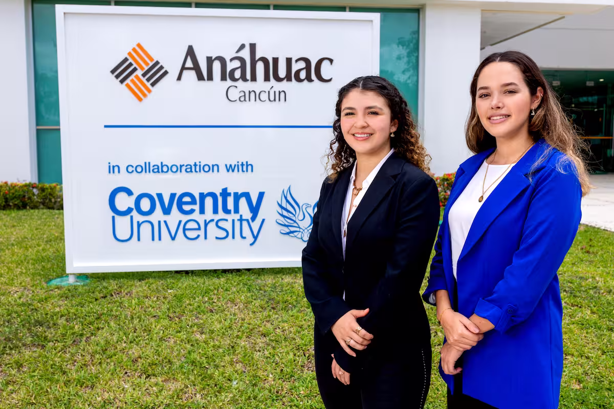 Dos mujeres jóvenes con vestimenta formal sonríen frente a un letrero de la Universidad Anáhuac Cancún, que destaca su colaboración con Coventry University. El fondo muestra un edificio moderno con áreas verdes. Dos mujeres jóvenes con vestimenta formal sonríen frente a un letrero de la Universidad Anáhuac Cancún, que destaca su colaboración con Coventry University. El fondo muestra un edificio moderno con áreas verdes.