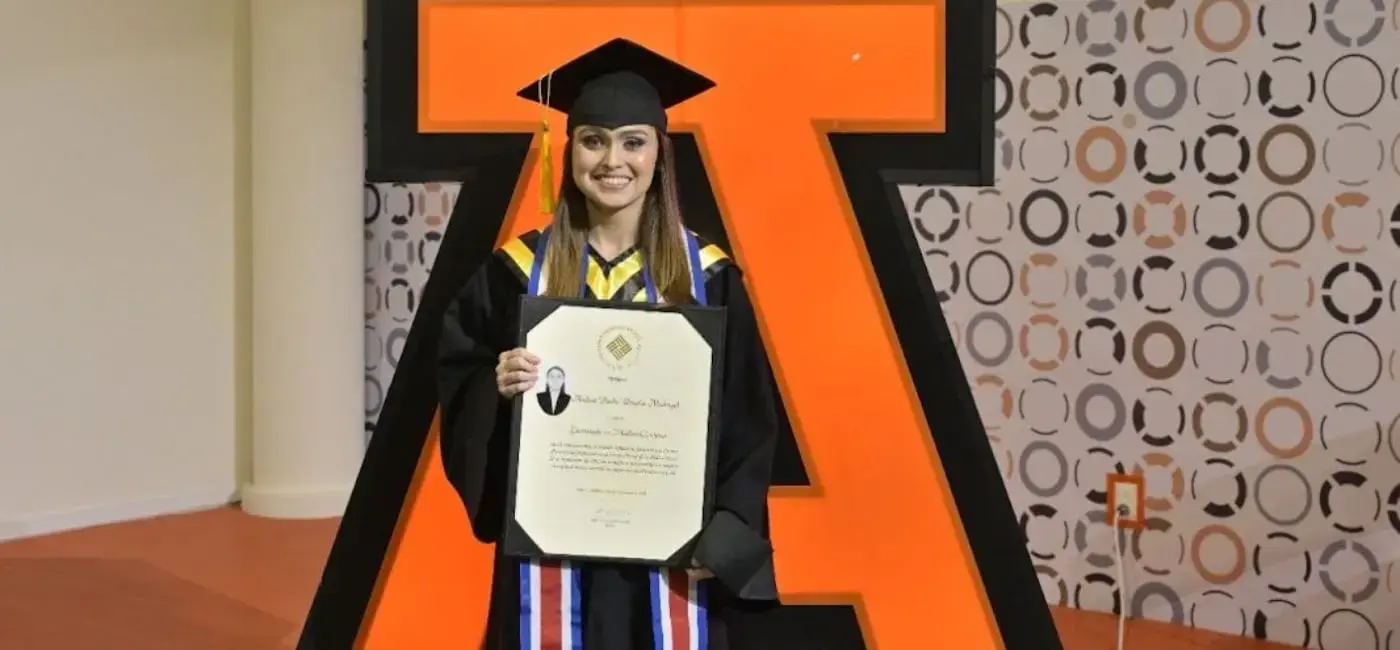 Una graduada sonriendo en toga y birrete sostiene su diploma frente a una gran letra 'A' naranja, logotipo de la universidad.