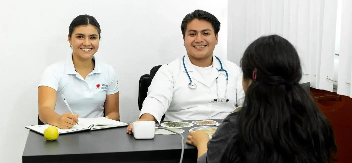Dos estudiantes de nutrición y salud sonriendo en una mesa de consulta práctica, con una libreta, monitor de presión arterial y fichas de alimentos, frente a una persona de espaldas siendo atendida.