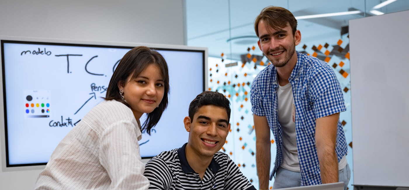 Tres estudiantes universitarios sonrientes colaboran en equipo frente a una pantalla digital con notas escritas, en un espacio moderno y multicultural.