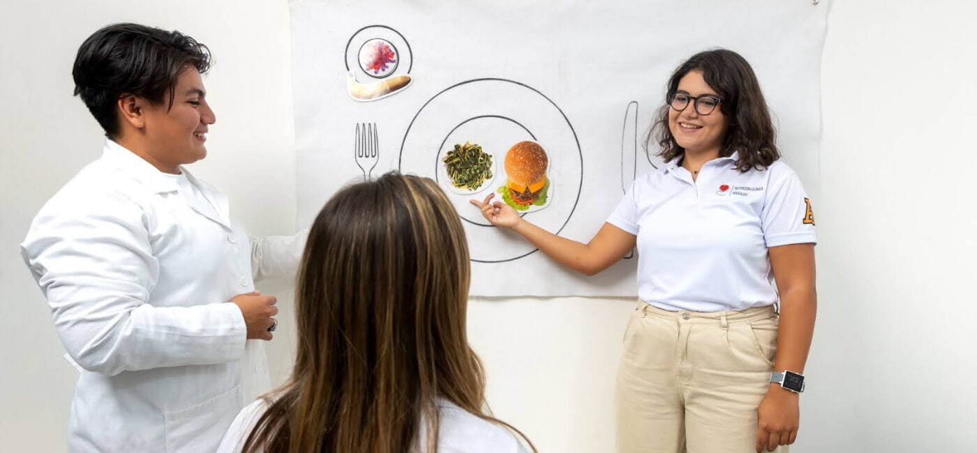 Dos estudiantes de nutrición dando una presentación a una compañera utilizando un cartel en la pared con el dibujo de un plato y recortes de alimentos.