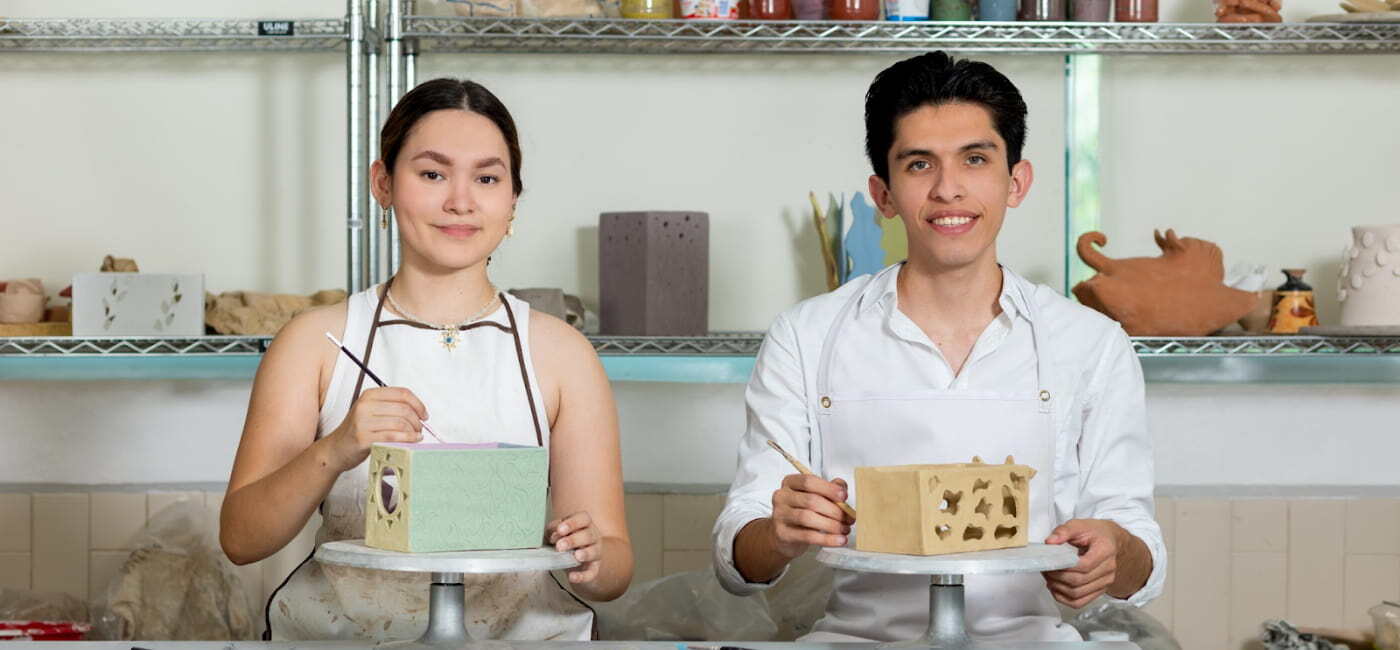 Dos estudiantes de Diseño Industrial, un hombre y una mujer, vestidos con mandiles protectores, trabajan en el diseño y decoración de piezas de cerámica o prototipos cuadrados sobre tornos de alfarería.