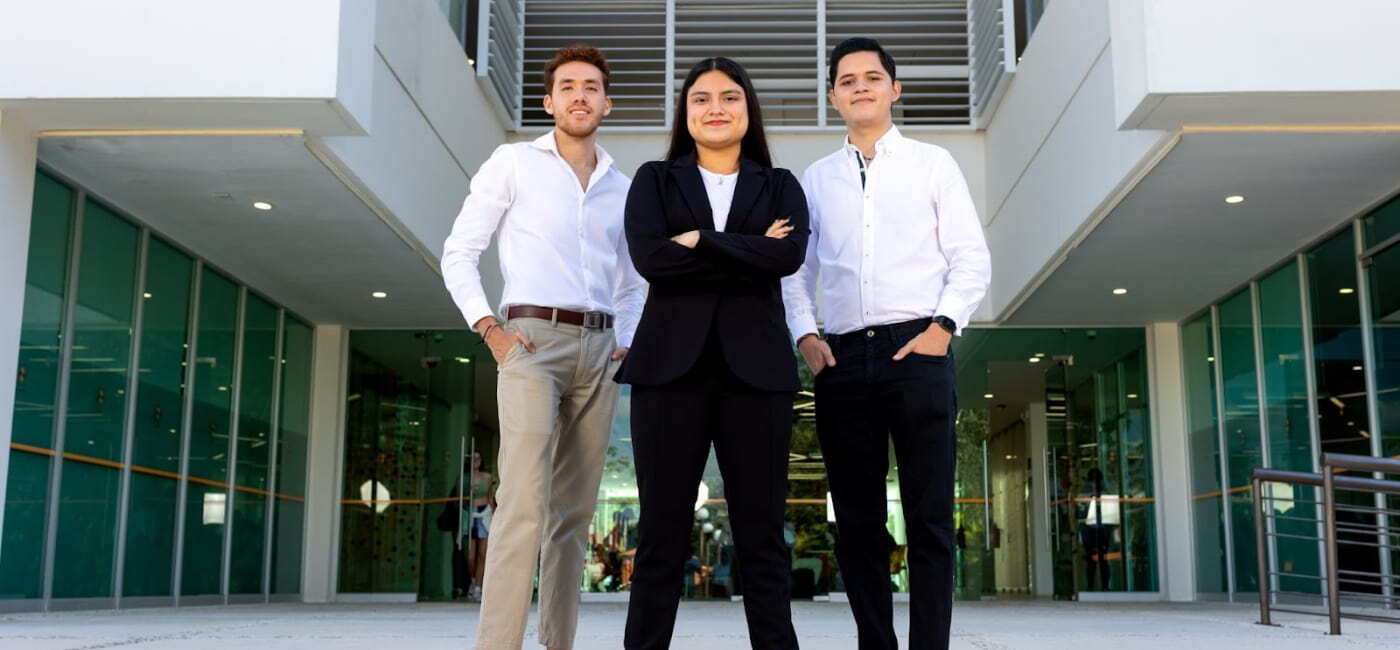 Tres estudiantes universitarios con vestimenta de negocios profesional posan con confianza frente a la fachada moderna de un edificio del campus universitario.