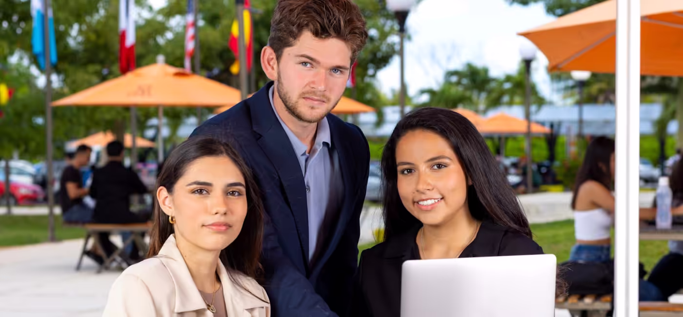 Tres estudiantes universitarios posan mirando a la cámara al aire libre en el campus, trabajando con una laptop en una zona de convivencia con sombrillas naranjas y banderas internacionales al fondo.