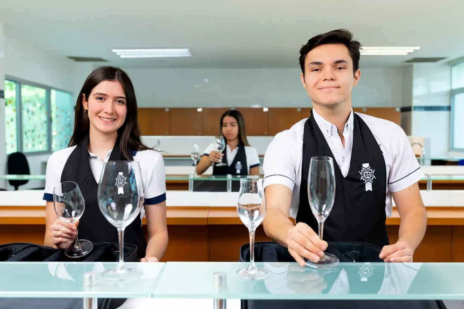 Dos estudiantes, un hombre y una mujer, posan sonrientes en un laboratorio de cata sosteniendo copas de cristal. Visten uniformes blancos con chalecos negros que llevan un emblema institucional.