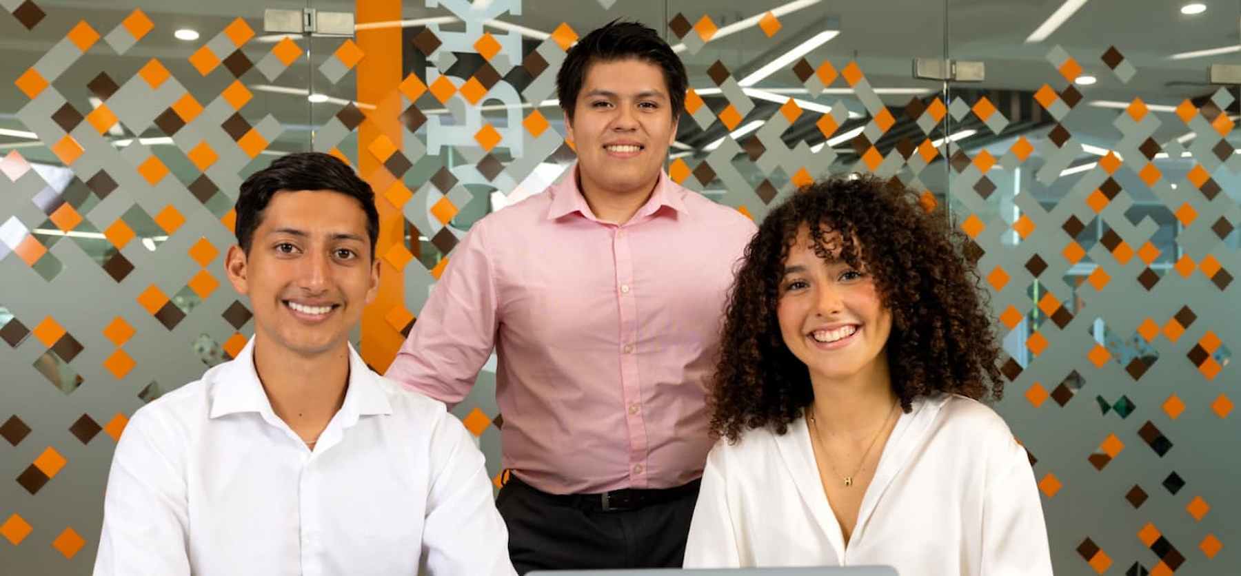 Tres estudiantes universitarios sonriendo frente a una pared de cristal decorada, representando el trabajo en equipo y el ambiente académico moderno.