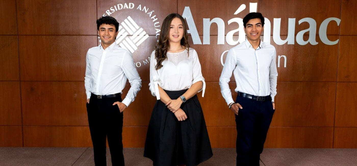 Tres estudiantes con vestimenta formal posan frente a una pared de madera con el logotipo de la Universidad Anáhuac.