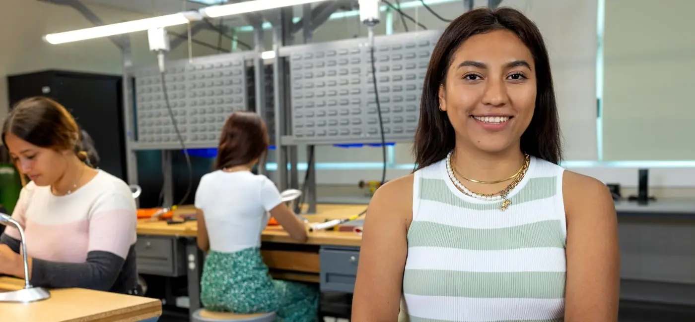 Una estudiante universitaria sonriendo en primer plano dentro de un laboratorio técnico, mientras sus compañeras trabajan concentradas en las mesas de trabajo con equipo especializado al fondo.