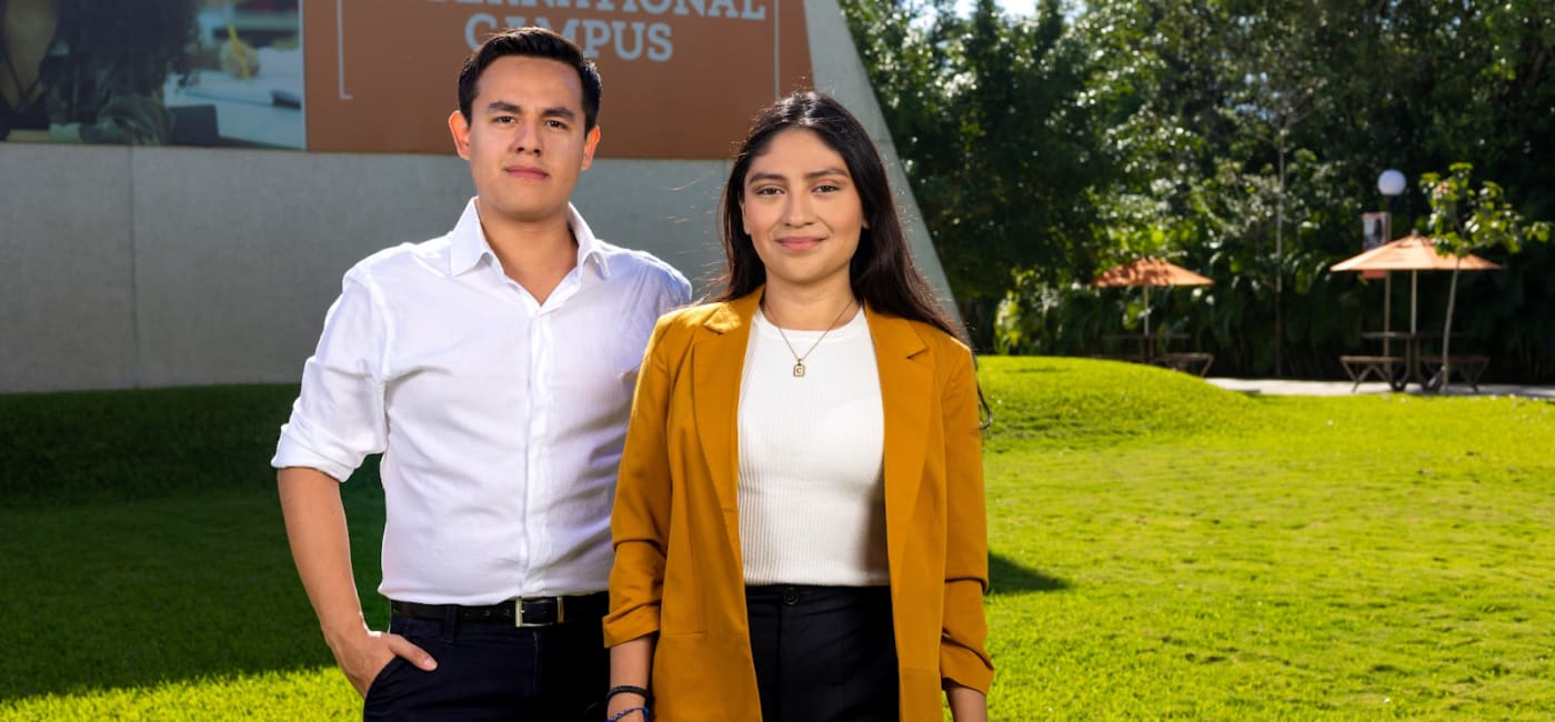 Dos estudiantes o jóvenes profesionales, un hombre y una mujer, posando al aire libre en un campus universitario con césped verde y un letrero institucional detrás. El hombre lleva una camisa blanca y pantalones oscuros, y la mujer un blazer color mostaza sobre un top blanco, simbolizando el Liderazgo y la Gestión Estratégica Internacional de la Universidad Anáhuac Cancún.