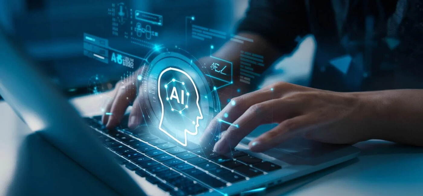 Primer plano de unas manos escribiendo en el teclado de una laptop en un ambiente oscuro, con un holograma futurista azul brillante superpuesto que muestra un perfil humano con las letras 'AI' y elementos de interfaz tecnológica.