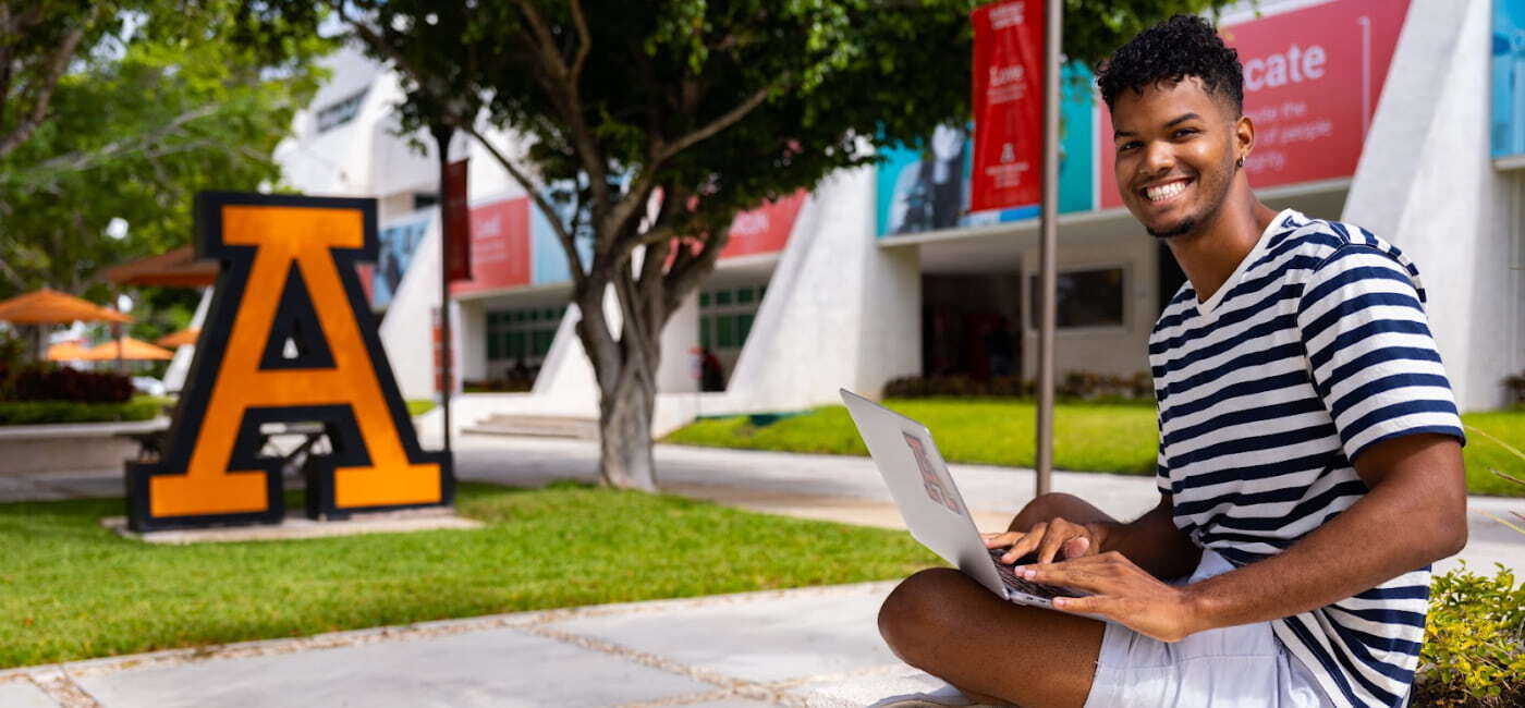 Estudiante universitario sonriente sentado en el campus con una laptop, junto a una escultura de la letra A gigante, rodeado de árboles y edificios institucionales.