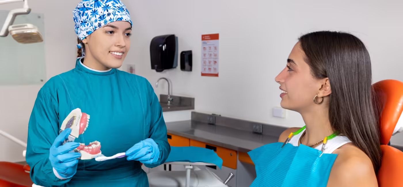 Una estudiante de odontología con uniforme clínico azul sonríe mientras le explica a una paciente en el sillón dental la técnica correcta de higiene, utilizando un modelo anatómico de dentadura y un cepillo en una clínica universitaria.